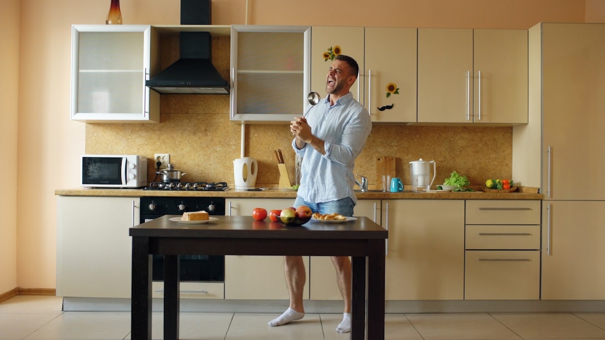 Attractive young funny man dancing and singing with ladle while cooking breakfast in the kitchen at home in the morining